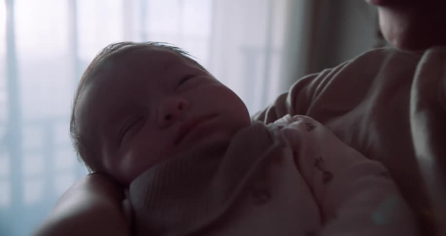 A sleeping newborn is cradled in a caregivers arms indoors softly lit by daylight filtering through sheer curtains conveying a quiet intimate moment