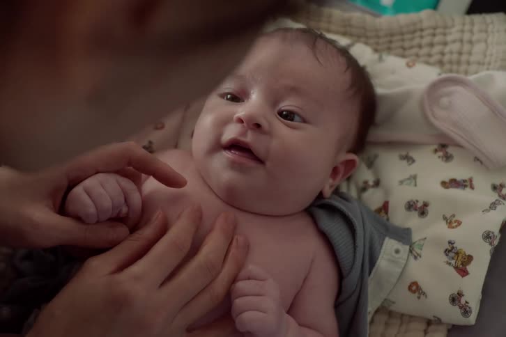 A wideeyed baby lies on a patterned blanket gently grasping a caregivers finger while being tenderly supported with a comforting touch