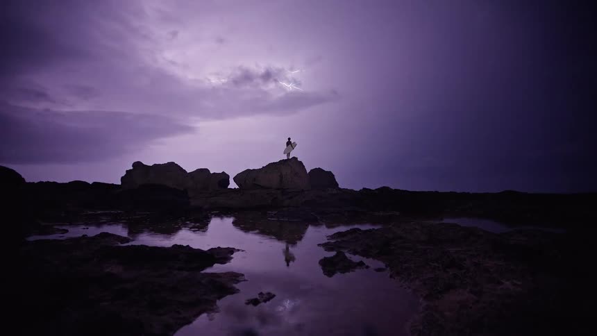 Lightning illuminates the night as the water reflects the person standing on a rock formation and holding a surfboard