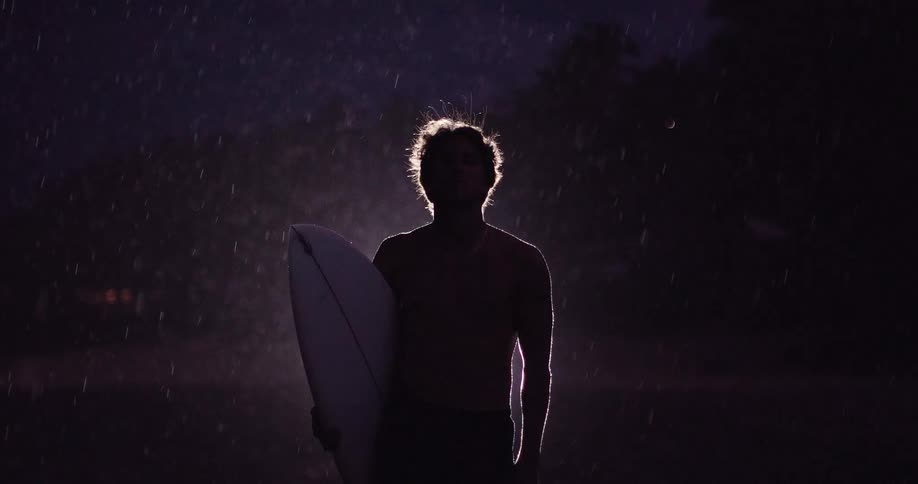 A man holds a surfboard while standing in a lightning storm at night