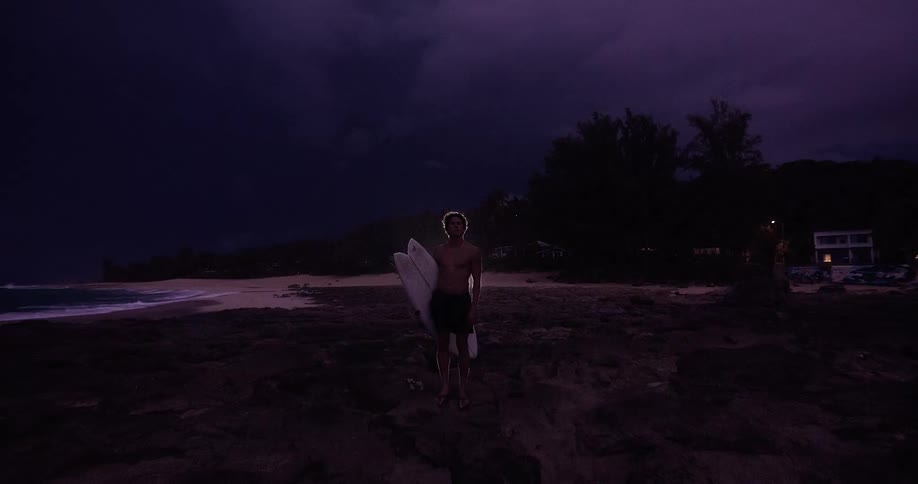 A man stands on the beach holding surfboards in a nighttime thunderstorm