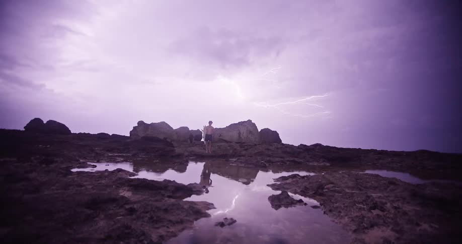 A man stands on a rocky tide pool landscape under a dark purple sky as a bright bolt of lightning illuminates the shore and reflects in the water