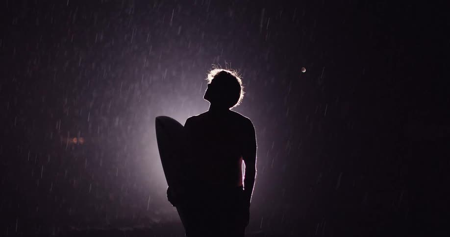 Silhouetted person looks upward holding a surfboard under rain with strong backlight