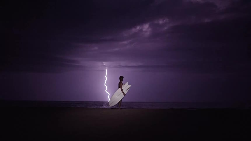 An adult holds a white surfboard while standing on an ocean coast during a lightning storm