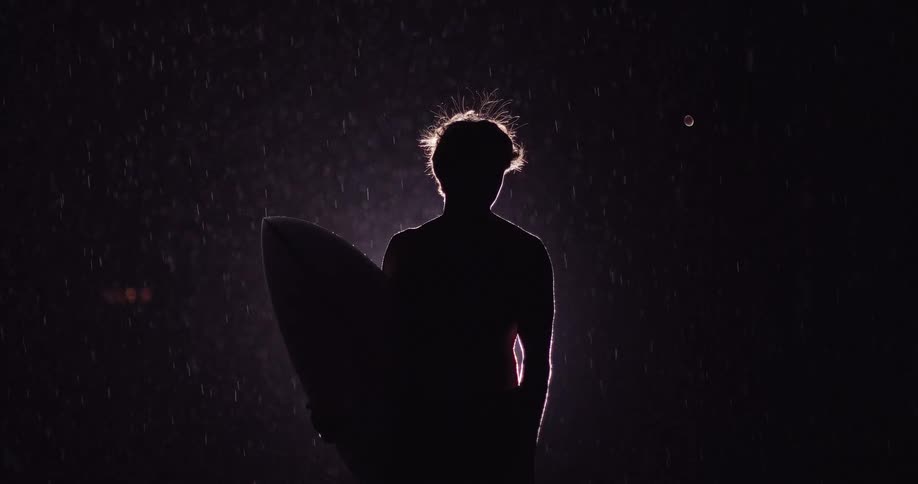 Silhouetted person holds a surfboard under rain in a dark setting with backlight