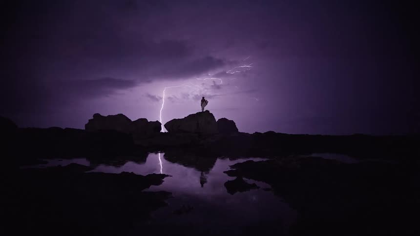 A lone surfer stands on dark rocks holding a surfboard as lightning streaks across a stormy night sky briefly illuminating the rugged shoreline
