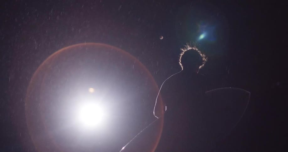 A man carries a surfboard while walking outdoors in a rainstorm at night