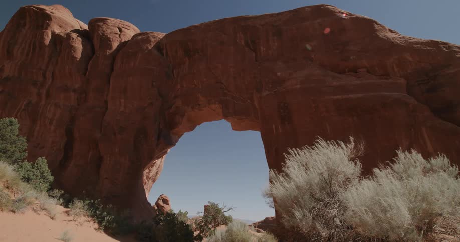 A massive natural sandstone arch towers over a desert landscape framed by a clear blue sky and sparse green shrubsthe camera captures a lowangle view of the rugged red rock formation highlighting its weathered texture and the bright sunlight hitting