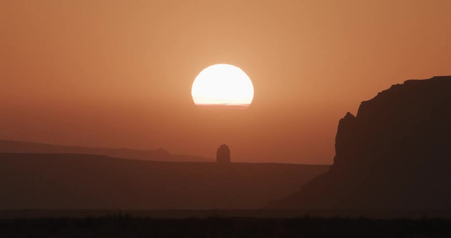Sun sets while mountains and rock formations stand silhouetted against a hazy red sky