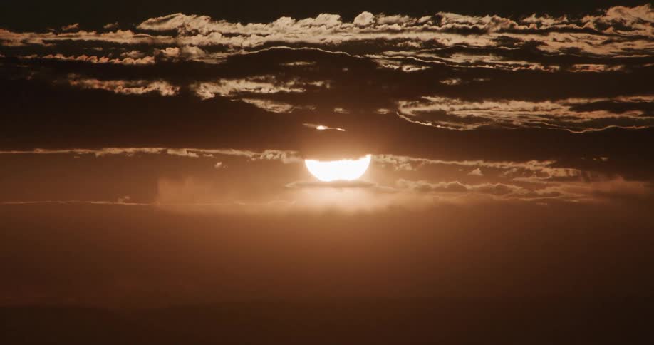The sun sets behind a layer of dark horizontal clouds over a vast desert casting a deep orange glow across the hazy atmosphere and silhouetting the distant mountain ridges on the horizon as the light fades
