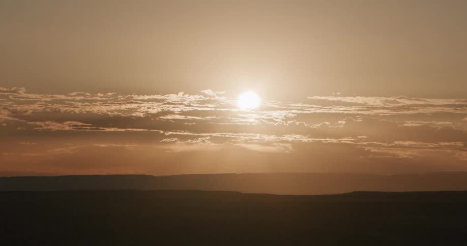 Passing clouds partially obscure the sun shining from a red sky onto hilly terrain on a hazy day
