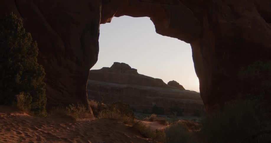 A view through a large natural rock arch reveals desert buttes and ridges under a pale sky during the golden hour as soft light illuminates the sandy ground and sparse desert plants