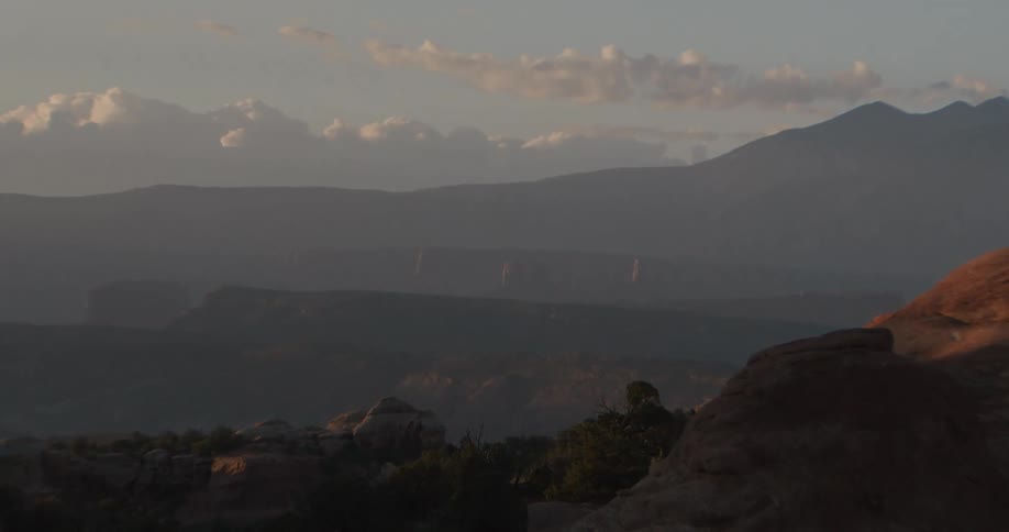 Vast desert landscape with layered rock formations and distant blue mountains under a pale sky during the late afternoon