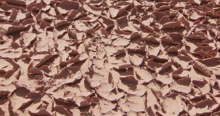 The camera slowly zooms in on a vast expanse of parched reddishbrown earth that has dried into curled petallike flakesthe highangle shot emphasizes the repetitive pattern of the cracked ground under bright direct sunlight