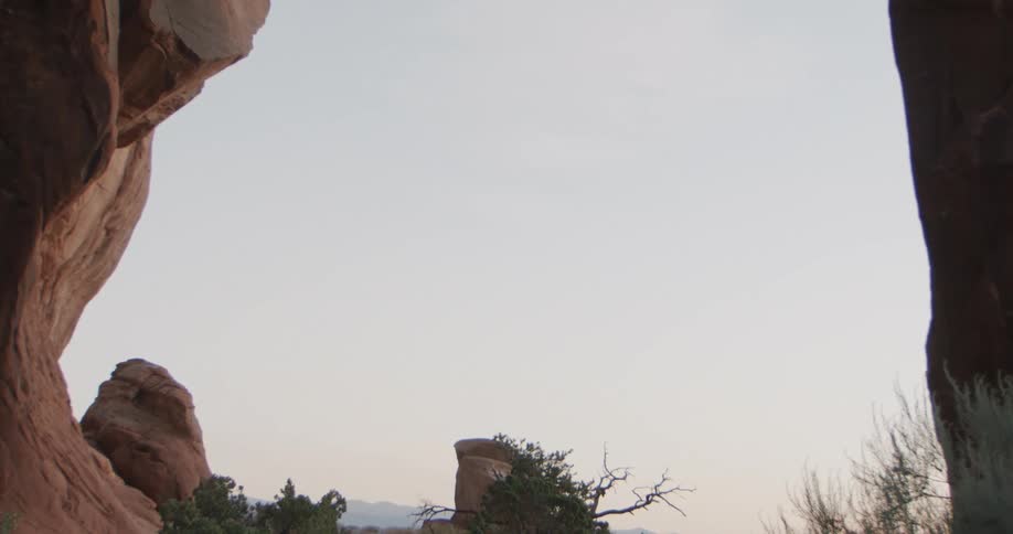 A camera goes from the base of a massive red sandstone arch in a desert moving from the shaded ground and desert vegetation to focus on the bright clear sky framed by the stone opening