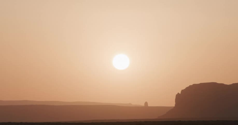A hazy sun sets over a vast desert plain with a rounded rock formation in the foreground and distant mountains fading into the warm golden horizon