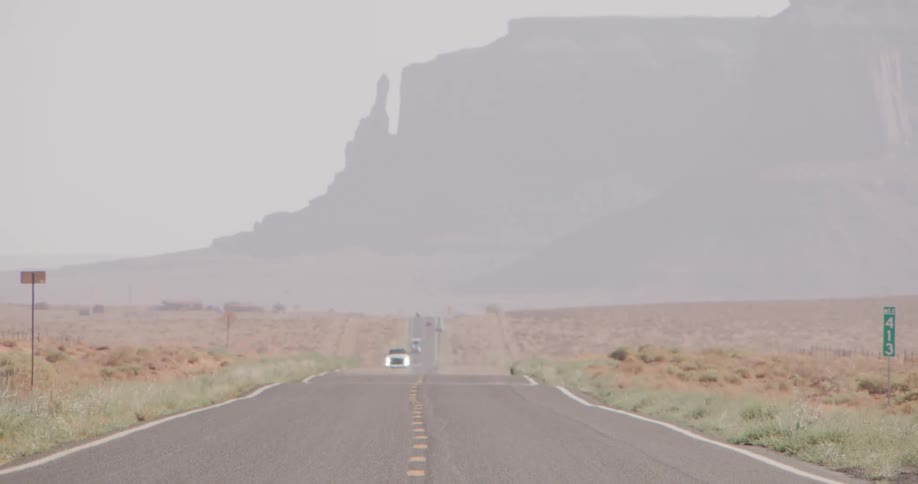 A lone car drives down an empty desert highway toward distant mesalike mountains shrouded in pale haze