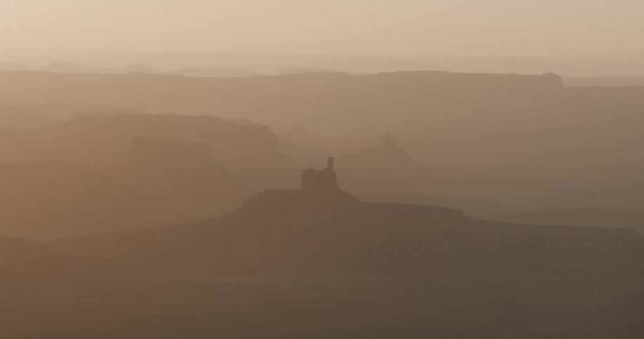 The camera slowly pans across a hazy sundrenched canyon landscape filled with silhouetted rock formationsas the lens moves the warm golden light reveals layers of mist settled between the deep ridges and towering buttes