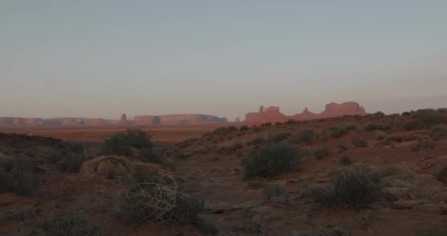 Low desert shrubs frame a rocky foreground opening toward distant mesas and buttes glowing softly in the fading light