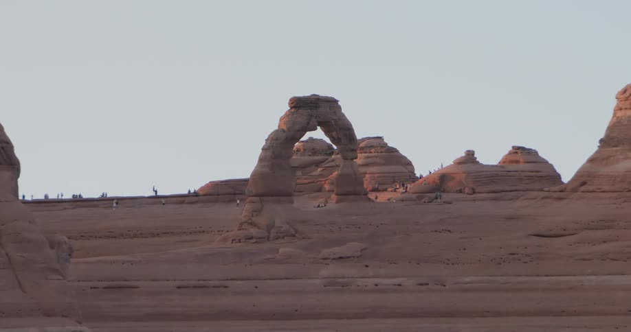 A wide shot zooms into a desert landscape during sunset showing several small tourists standing around the massive sandstone formation and distant desert ridges under a clear sky