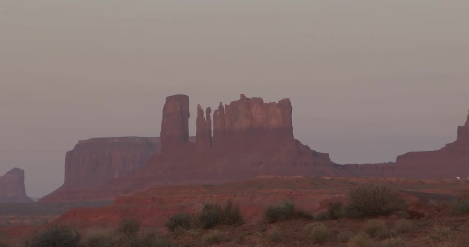 Massive sandstone buttes and rock towers rise from a flat desert floor under a soft purple and orange sky during a quiet sunset