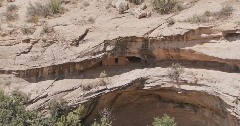 An aerial camera zooms in on a sundrenched desert canyon wall to reveal ancient stone ruins the structures are built deep within a natural rock alcove featuring a small rectangular window and stacked masonry