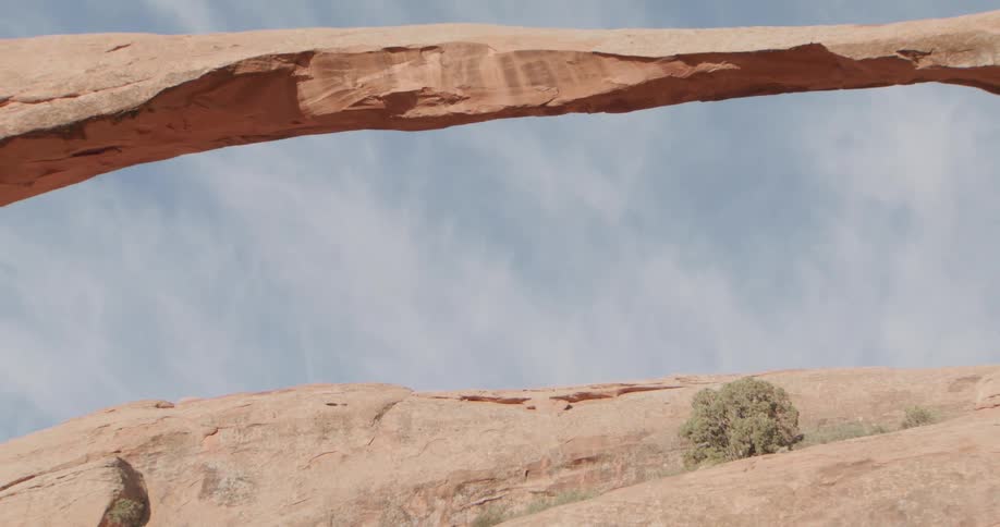 The camera tilts down from a pale wispy blue sky to reveal a thin natural sandstone arch spanning across a desert landscapeas the view pulls back the immense scale of the rock formation is shown against a backdrop of rugged sundrenched cliffs