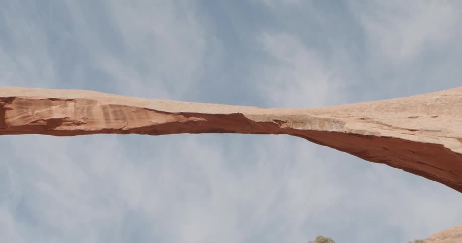 A natural stone arch spans across a desert canyon beneath a lightly clouded blue sky
