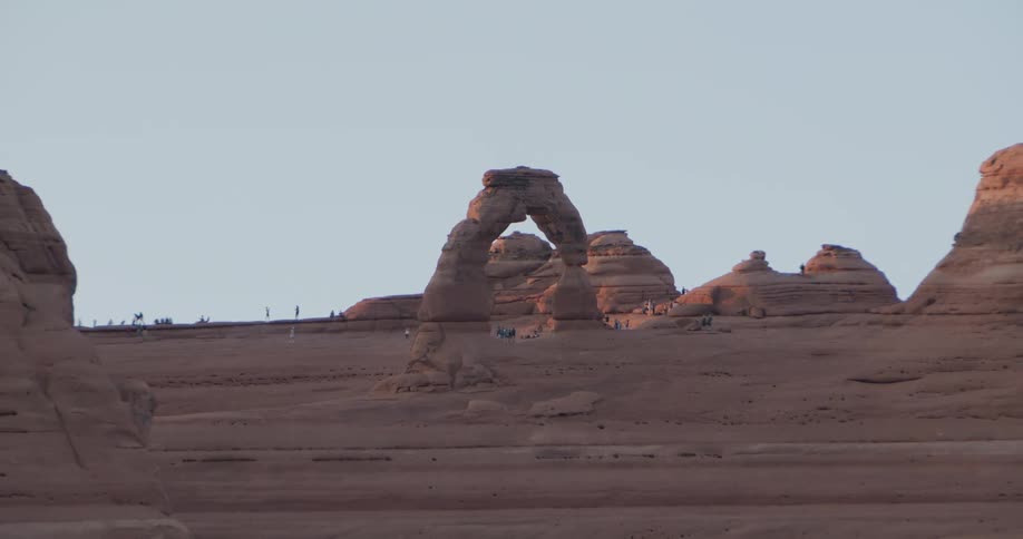 The video shows a wide tracking shot of delicate arch in arches national park utah as the camera pulls back to reveal the surrounding red rock formations numerous tourists are visible walking and standing near the base of the arch and along the adjacen