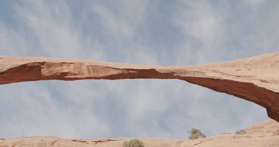 Desert plants grow under a narrow stone arch and near the base of a rocky hill on a cloudy day