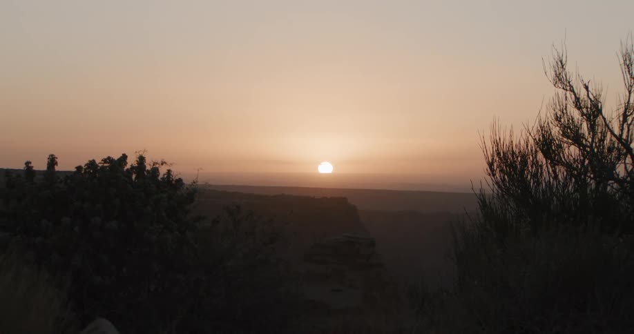 The video features a slow zoom out from a bright sun setting over a vast hazy desert canyonas the camera retreats dark silhouettes of rugged terrain and sparse vegetation emerge in the foreground against the glowing orange sky