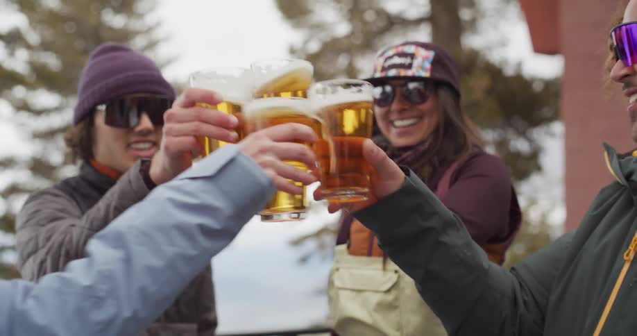 People wearing cold weather wear clink beer glasses while standing on a balcony near the water during the daytime