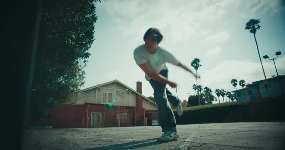 A young man in a white tshirt and loose pants walks across a sunny paved lot while carrying his skateboard behind him tall palm trees and suburban houses under a bright sky create a classic southern california atmosphere