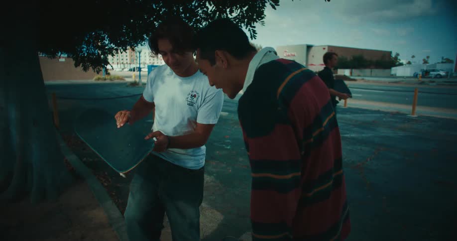 Two young men are focused on a skateboard one man in a white tshirt is holding the board and pointing at the wheels or the underside while the man in the striped shirt leans in to inspect it their interaction creates a stationary point of interest tha