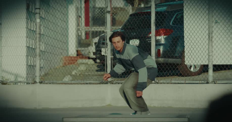 A young man in a striped longsleeve polo shirt focuses with his eyes closed before skating forward he successfully performs a trick onto a concrete ledge and rolls away with a celebratory smile