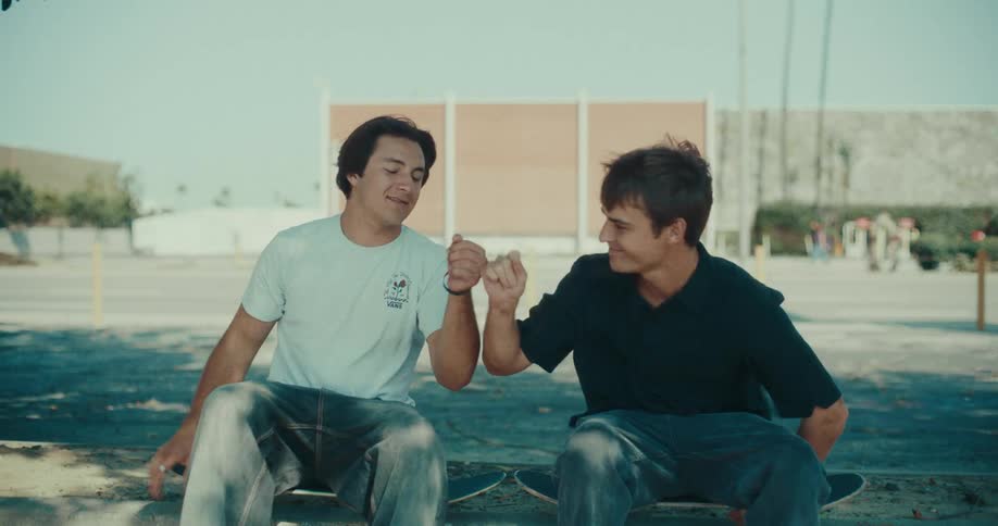 Two young men sit on their skateboards in a parking lot share a fist bump while smiling and then stand up to walk away with their boards in hand