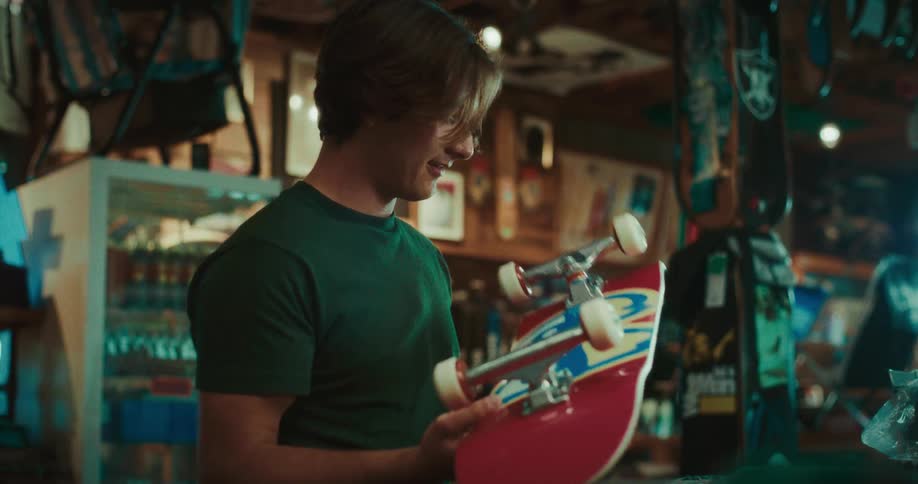 Inside a warmly lit skate shop a young man smiles as he inspects a red skateboard handed to him sharing the happy moment with a friend