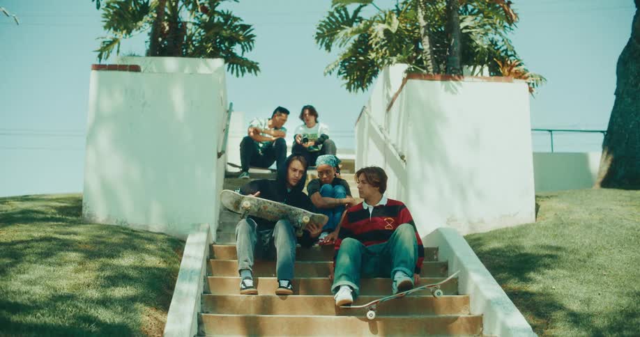 A group of teenage friends sitting on a stairway at midday holding skateboards as they talk together
