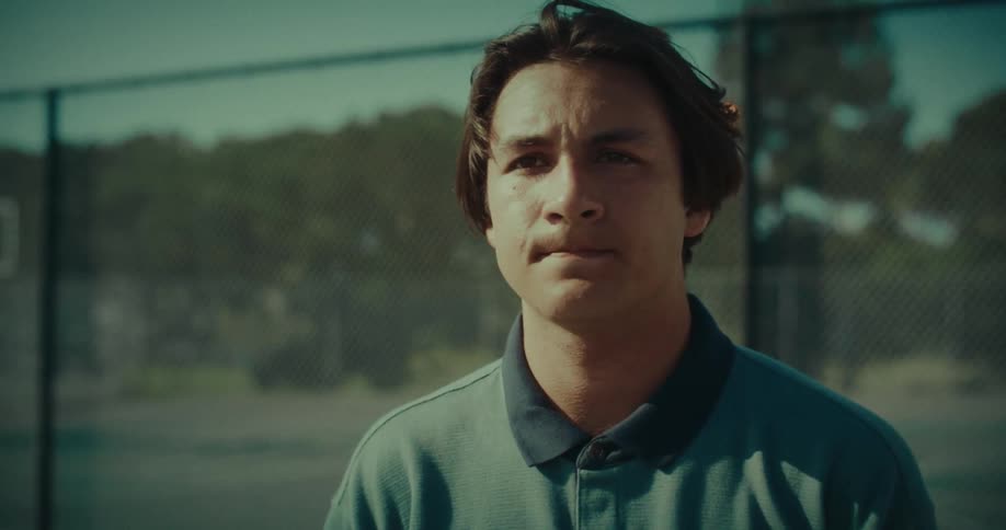 Teenage boy standing near a fence and he pushes back his hair with his hand and moves away
