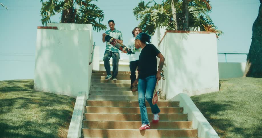 A group of young people carrying skateboards stands up and begins walking down a wide set of outdoor concrete stairsthe diverse group of friends moves toward the camera and eventually exits the frame on a sunny day