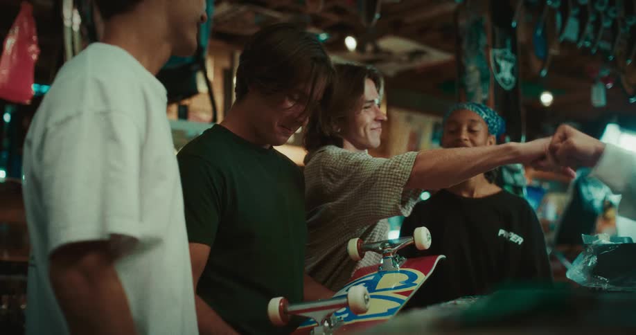 Small group of teenagers stand inside a shop as a boy holds a skateboard upside down and spins the wheels