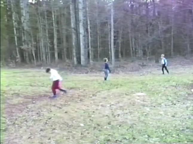Three children run across a grassy clearing toward a wooded area before slowing down and stopping near the edge of the forest