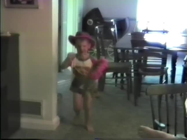 A little boy with a pink cowboy hat is riding a toy stick horse through a house