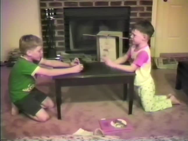 Two boys playing with a paper football on a table in the living room as a little girl walks by