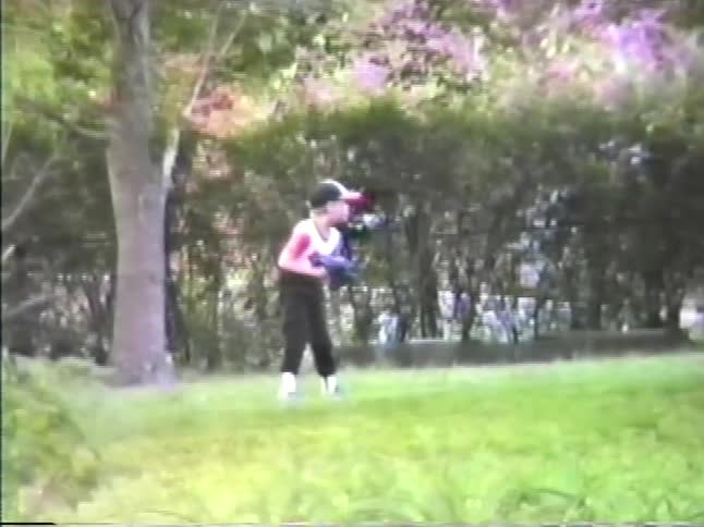 A boy in a public park at midday wearing a baseball hat and gloves throwing a baseball across the field
