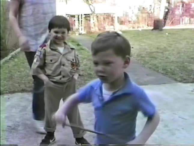A young boy wearing a tan scout uniform stands on a concrete path while another boy in a blue shirt runs nearby in a grassy yard