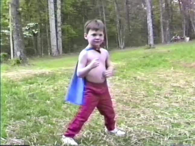 A young shirtless boy in red pants and a blue cape strikes several heroic poses in a grassy backyard while another child passes in the background near a line of trees