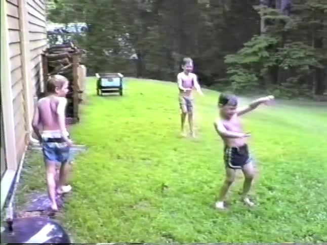 Three young boys in swim trunks laugh and run through a water sprinklers spray on a grassy lawn next to a house and a wooded area