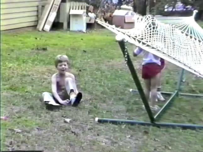 A young boy with no shirt runs and cheers in a grassy yard while other children play near a white rope hammock on a summer day