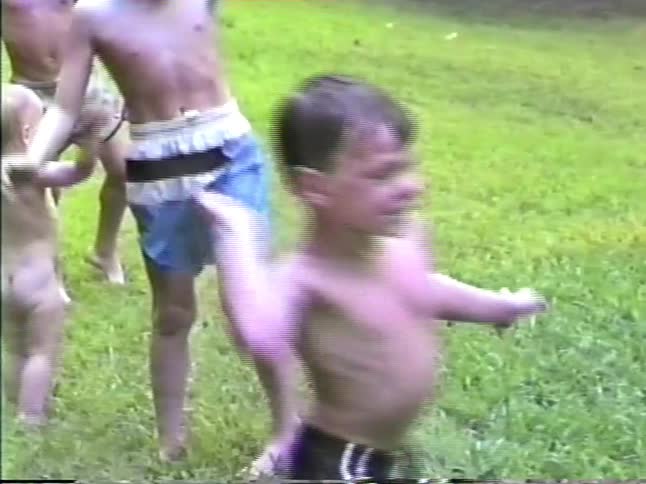 Young boys in swim trunks run and dance through a water spray from an offscreen sprinkler in a grassy backyard bordered by trees while a toddler watches near by